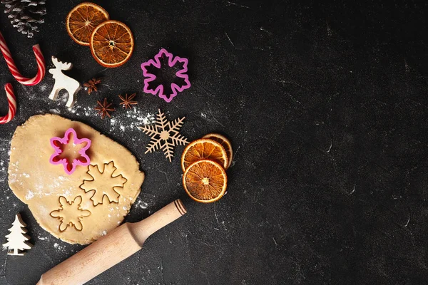Cookie dough with Christmas cookie cutters, Christmas ornaments. Making gingerbread cookies for winter holidays. Dry orange slices,  candy cones and wooden New Year decoration on the black background.