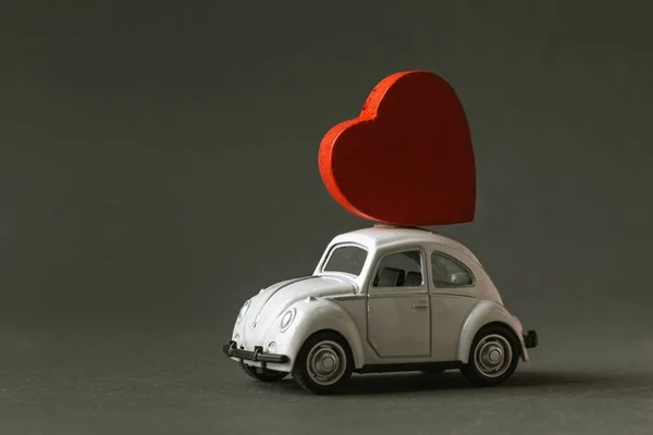 Retro toy car delivering wooden red heart at the roof for valentine\'s day at the blurred dark grey background.