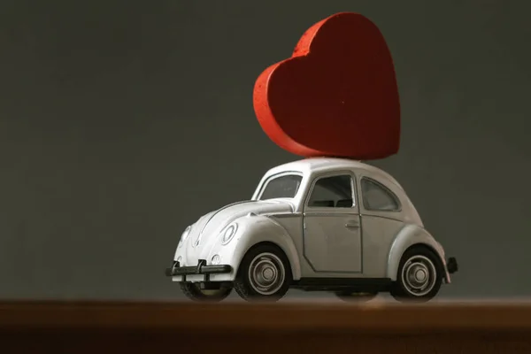 Retro toy car delivering wooden red heart at the roof for valentine\'s day at the blurred dark grey background.