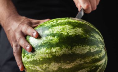 Adamın biri siyah arka planda büyük bir karpuzu bıçakla kesiyor. Erkeklerin ellerinin karpuz tutarken yakın plan çekimleri.