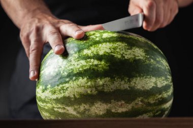 Adamın biri siyah arka planda büyük bir karpuzu bıçakla kesiyor. Erkeklerin ellerinin karpuz tutarken yakın plan çekimleri.