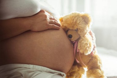 Hamile kadın büyük çıplak karnına dokunuyor ve oyuncak ayıyı pembe kurdeleyle tutuyor. Bebek kız beklentisi. Sevgi ve ilgi. Kapat.