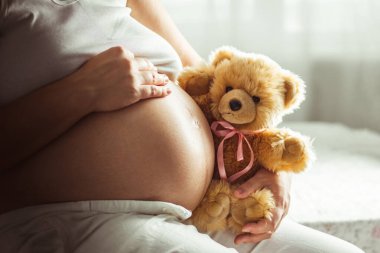 Hamile kadın büyük çıplak karnına dokunuyor ve oyuncak ayıyı pembe kurdeleyle tutuyor. Bebek kız beklentisi. Sevgi ve ilgi. Kapat.