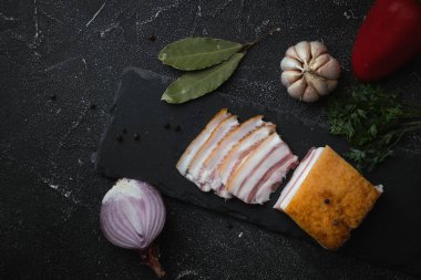 Saha yaprağı, sarımsak ve soğanla süslenmiş siyah beton arka planda dilimlenmiş tuzlanmış domuz pastırması. Sağlıklı yemek olarak domuz eti. Üst Manzara. Geleneksel Ukrayna yemekleri