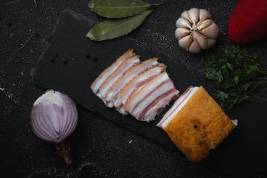 Saha yaprağı, sarımsak ve soğanla süslenmiş siyah beton arka planda dilimlenmiş tuzlanmış domuz pastırması. Sağlıklı yemek olarak domuz eti. Üst Manzara. Geleneksel Ukrayna yemekleri