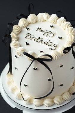 Zarif Lambeth tarzı doğum günü pastası krem rengi krem peynirli süslemeli ve üstünde siyah mutlu yıllar mesajı olan. Siyah arkaplan