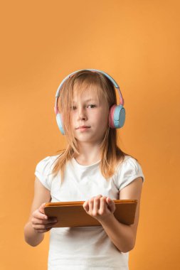 Büyük kulaklık takan ve bej arka planda tablet tutan sevimli sarışın kız. Çalışma veya oyun konsepti için tablet bilgisayar kullanılıyor.