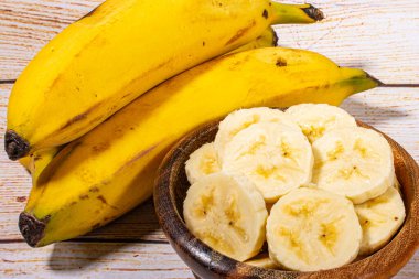 Muz, tahta bir kasede dilimler halinde kesilmiş ve arka planda muzlar açık ahşap bir arka planda hasır bir sepette..