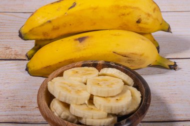 Muz, tahta bir kasede dilimler halinde kesilmiş ve arka planda muzlar açık ahşap bir arka planda hasır bir sepette..