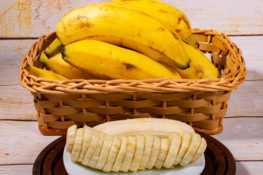 Tahta tepside beyaz bir tabakta dilimlenmiş muz ve arkasında muz olan hasır bir sepet.