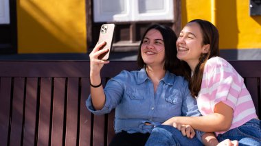 Güzel lezbiyen çift sokakta birbirlerini seviyorlar, telefonla fotoğraf çekiyorlar, gülüyorlar ve LGBT onur bayrağıyla yürüyorlar.