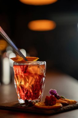 Kurutulmuş portakal dilimleri ve bardaki çiçeklerin yanında taş bardakta servis edilen eski moda kokteyl..