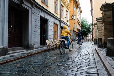Stockholm 'deki Gamla Stan' de kaldırımtaşlı caddede bisiklet süren insanlar var.