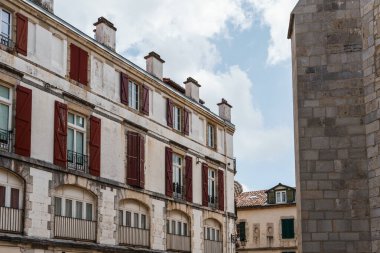 Görüntü, Fransa 'nın Bayonne şehrinde bulutlu bir gökyüzüne kurulmuş, beyaz cepheli ve koyu yeşil panjurlu geleneksel binaları gösteriyor..