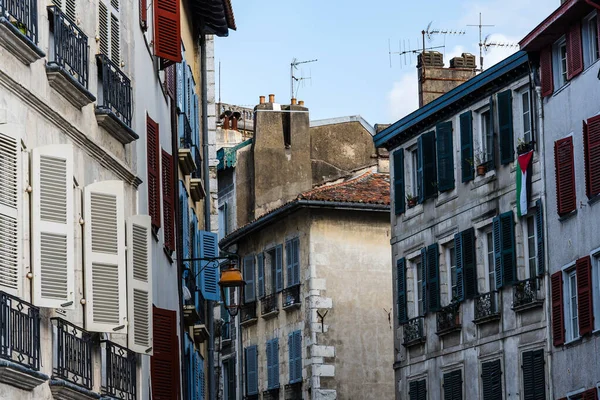 Görüntü, Fransa 'nın Bayonne şehrinde bulutlu bir gökyüzüne kurulmuş, beyaz cepheli ve koyu yeşil panjurlu geleneksel binaları gösteriyor..