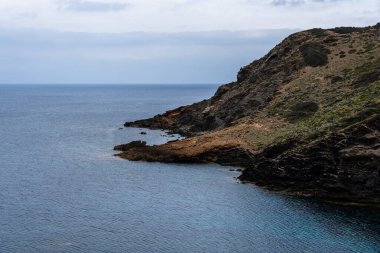 Görüntü, Menorca 'nın engebeli sahil şeridinin engebeli güzelliğini yansıtıyor. Yumuşak, bulutlu bir gökyüzünün altında berrak mavi denize düşen dramatik uçurumlar var..