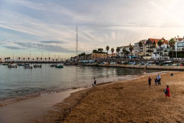 Cascais, Portekiz - 30 Ekim 2022: Güzel bir Cascais manzarası, Portekiz, kumlu bir sahil, limanda tekneler, ve gün batımında sahil boyunca binalar.