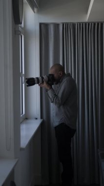 Pencere kenarındaki gözetleme fotoğraflarını çekmek için telefoto merceği kullanan kel bir erkek. Dikey Video, Kilitli