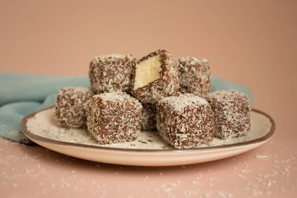 Traditional Australian cuisine, Lamington cakes with chocolate icing and coconut flakes on a ceramic plate on a coral pink background. Sponge cake baked goods.