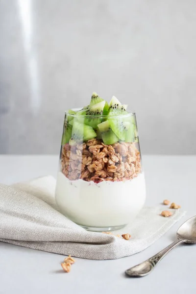 Nutritious healthy snack or breakfast. Homemade yogurt,sour cream or pudding with kiwi and granola in a glass on a gray background lit from a window. Light layered organic dessert with spoon and napkin. Concept for overlay on mockup,template,posters.