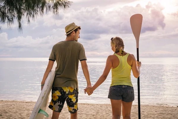 Pareja feliz con tabla de paddle en la playa al atardecer — Foto de Stock