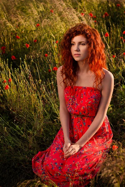 Red-haired girl sitting in the poppies meadow at sunset — Stock Photo, Image