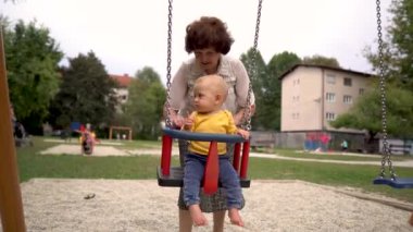 Bir büyükanne parkta salıncakta sallanan bir bebeği nazikçe sallıyor. 4K