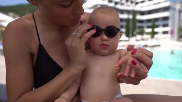 Mamãe colocando óculos de sol pretos em seu bebê menino em uma piscina 4K — Vídeo de Stock