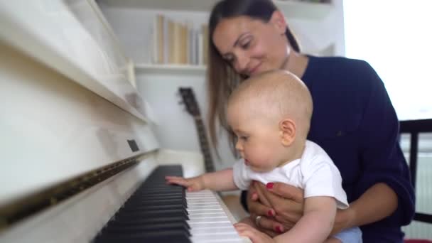 Een schattige baby jongen spelen een witte piano met zijn mama leren hem en houd hem in haar schoot 4K — Stockvideo