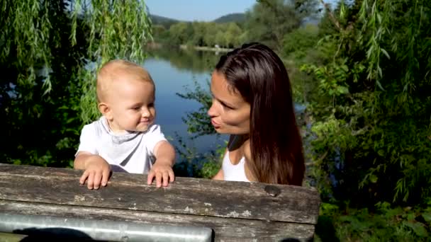 一个可爱的母亲和一个婴儿在一个漂亮的湖畔的长椅上。妈妈亲了个小宝宝4K — 图库视频影像