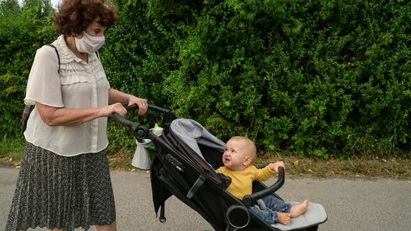 A grandmother with a medical mask pushing an upset baby boy in a stroller on a street. Covid-19 separating families.