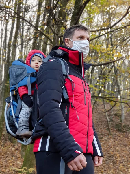 Tıbbi koruyucu maske takan bir baba ve ormanda bir bebek taşıyıcısı.