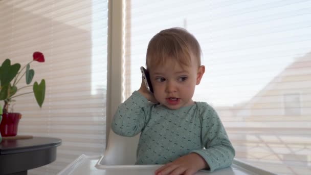 Um bebé giro a falar num telemóvel numa cadeira de bebé. 4K — Vídeo de Stock