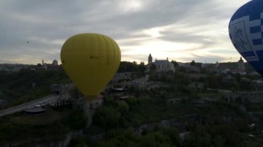 Renkli bir sıcak hava balonunun insansız hava aracı yeşil bir parkın üzerinde uçarken görüntüsü ve yazın gündoğumunda küçük bir Avrupa şehrinde bir nehir.