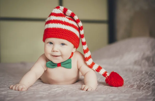 Örgü örmüş Noel Baba şapkalı mavi gözlü bir bebek karnının üzerine uzanmış, yukarı bakıyor ve gülümsüyor..