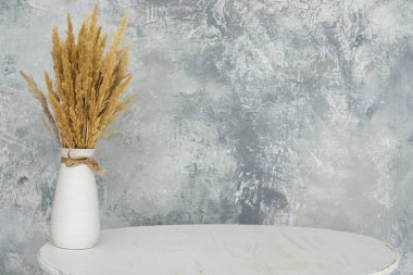 Gri bir duvara karşı beyaz bir masanın üzerinde buğday gibi duran beyaz bir vazo. Bir ürünün reklamını yapmak için bir yer var..