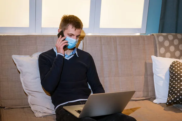 young guy talking on phone wearing medical mask sitting on the couch at home