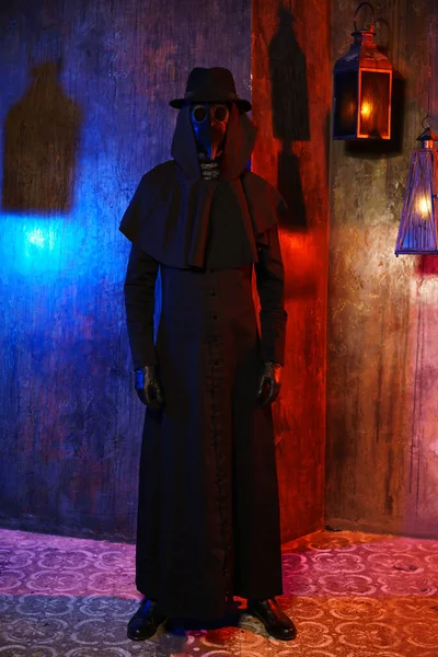 Portrait of a plague doctor in a black leather mask against a dark wall with lanterns.