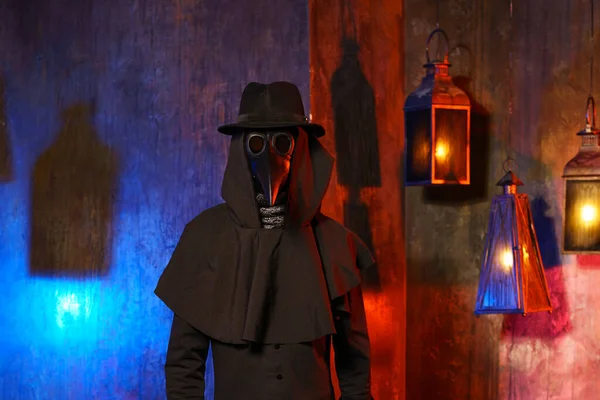 Portrait of a plague doctor in a black leather mask against a dark wall with lanterns.