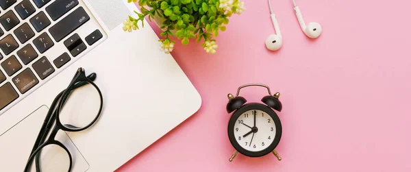 Alarm clock and glasses, white wired headphones and laptop on pink background.