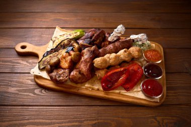 Assorted grilled meats with vegetables and dipping sauces served on wooden board for barbecue or sharing meal concept.