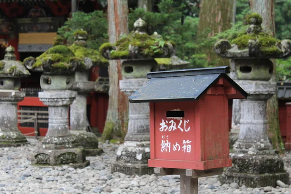 Red collection box at Japanese Shinto Buddhist shrine temple with stone lanterns in background. Translation: \