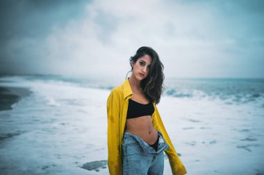 Okyanusun karşısındaki sahilde poz veren genç bir kadın, suyun içinde duruyor.