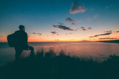 Sırt çantalı bir adam gün batımında nehre bakıyor.