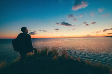 Sırt çantalı bir adam gün batımında nehre bakıyor.