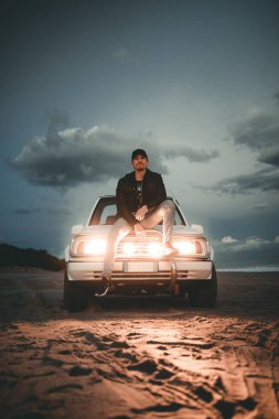 Genç adam akşam plajda araba kaputunda oturuyor. 