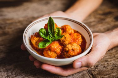 Elleri kasede, ahşap masada domates soslu köfte tutuyor.