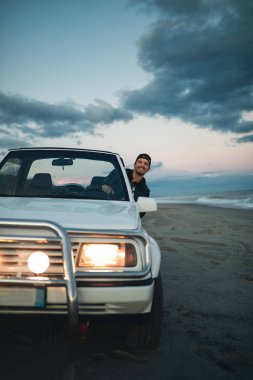 Genç adam deniz kıyısında arabada oturuyor.