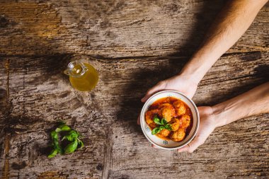 Elleri kasede, ahşap masada domates soslu köfte tutuyor.