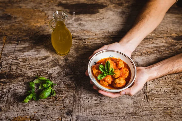 Elleri kasede, ahşap masada domates soslu köfte tutuyor.